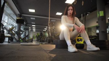 Spor giyim ve yüksek topuklu ayakkabı giyen bir kadın spor salonundaki bankta dinleniyor. Yanında bavulları ve bavulları olan bir çantası var. Flaş fotoğrafçılık yasaktır