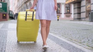 Şehrin arka planında kaldırım taşları üzerinde yürüyen, sarı bir bavulu çeken genç bir kadının seksi bacakları görünüyor. Bagajı olan bir turist kadın. Parlak sarı seyahat valizi.