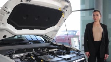 İş stili olan bir kadın açık bir arabanın kaputunun yanında duruyor. Araba tamiri. Araba servisi. Arabayı içeriden gösteriyor.