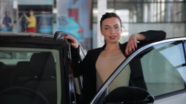 Çekici genç bir iş kadını araba galerisinde açık bir arabaya yaslanıyor, araba galerisinde. İş kadını yeni bir araba seçer.