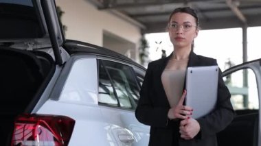 Bir araba galerisinde açık bir arabanın yanında dizüstü bilgisayar tutan kendine güvenen genç bir kadın. Bir araba galerisinin kadın satış danışmanı, satış müdürü..