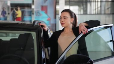 Çekici genç bir iş kadını araba galerisinde açık bir arabaya yaslanıyor, araba galerisinde. İş kadını yeni bir araba seçer.