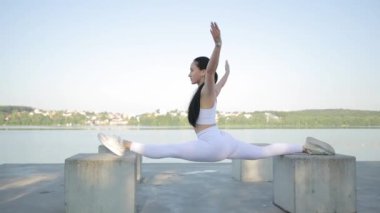 Beyaz eşofmanlı zayıf bir kadın göl kıyısında gökyüzüne karşı taşların üzerinde meditasyon yaparken ikiye ayrılıyor. Germe egzersizleri ve meditasyon. Yoga yapan konsantre bir kadın.