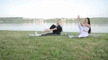 İki Avrupalı genç kadın bir şehir gölünün kıyısında otururken meditasyon yapıyorlar. Yoga pozları. Yoga sabah gölünün kıyısında temiz havada gökyüzünün arka planında.