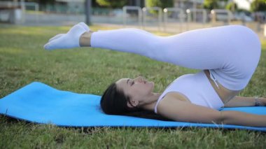 Sıcak bir yaz gününde yeşil bir alanda jimnastik minderinin üzerinde uzanırken esneme egzersizleri yapan sıska bir genç kadın. Temiz havada spor eğitimi.
