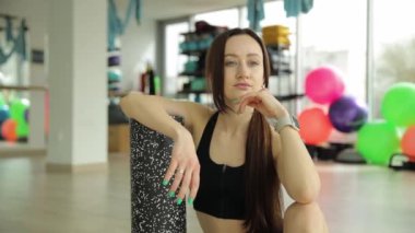 Spor salonunda oturan bir kadın köpük silindir ve yoga minderine yaslanıyor. Barışçıl ortam idmanlar arasında mola ya da meditasyon yapmayı ima eder.