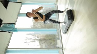 Bir kadın spor salonunda koşu bantları, makineler ve platformlar gibi farklı ekipmanlar kullanarak sıkı çalışır. Çömelme kuvveti ve dengeyi bir pencerenin önünde ayakta durarak gösterir.