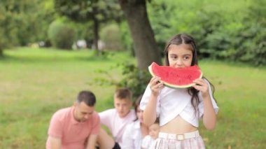 Küçük bir kız yaz parkında bir dilim karpuz yer, aile arka plandadır. Yüksek kalite 4k görüntü