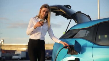 Bir kadın elektrikli arabasını şarj ederken telefonundaki güncellemeleri kontrol ediyor, elektrikli araçların ve ulaşım teknolojisinin kolaylığını vurguluyor
