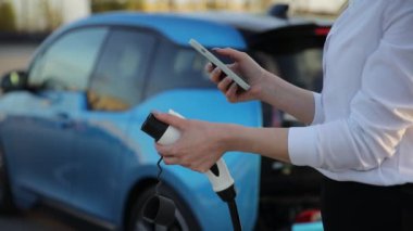 Elektrikli arabasının yanında duran bir kadın cep telefonuyla şarj ediyor. Araç kaputu açık, lastik ve tekerlek gibi otomotiv parçalarını gösteriyor.