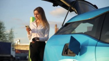 Bir kadın telefonunu elektrikli bir arabanın yanında kullanır, sonra da telefonuyla arabayı şarj eder. Sonra, mavi elektrikli bir arabanın yanında duruyor, hala telefonuyla.