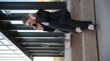 Takım elbiseli profesyonel bir kadın bankta oturur ve telefonda konuşur. Odaklanmış ve sohbete dalmış görünüyor.