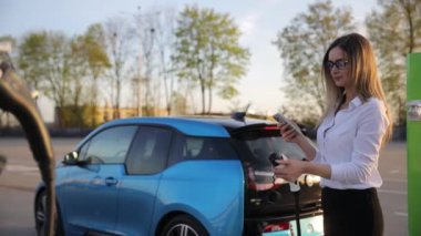 Beyaz, neşeli, genç bir kadın şehirde akıllı telefondan arıyor elektrikli arabasının şarj edilmesini bekliyor. Güzel mutlu iş kadını.