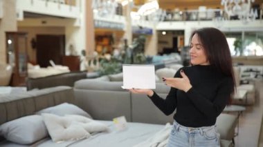 Bir kadın, bir mobilya mağazasında, elinde tablet, cep telefonuyla konuşuyor, bir kutu taşıyor, dizüstü bilgisayarını tutuyor, yoğun bir alışveriş ve iletişim gününü gösteriyor.