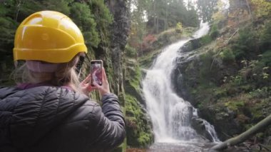 Sarı şapkalı bir kadın, doğa ve fotoğrafçılığa olan tutkusunu sergileyerek fevkalade bir şelalenin fotograflarını çekiyor.