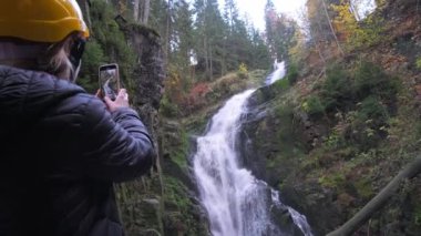 Bir kadın akıllı telefonuyla görkemli bir şelalenin güzelliğini yakalar. Başka bir insan bu anı paylaşıyor, doğayla olan bağlantısını gösteriyor.