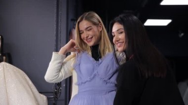 Gülümseyen ve poz veren iki mutlu kadın bir giyim mağazasında. Biri katmanlı saçlı ve uzun kirpikli mor bir elbise denerken, ikisi de gösterimde şık bir mor elbiseye hayran kalıyor.