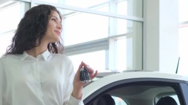 Bir kadın, binanın önüne park etmiş bir aracın önünde arabanın anahtarıyla jest yapıyor. Pencerenin yanında duruyor, otomotiv yarışması için hazır.