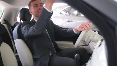 Takım elbiseli bir adam, bir motorlu aracın sürücü koltuğunda oturur ve araba sürmeye hazırlanırken dış tasarımı gösterir.