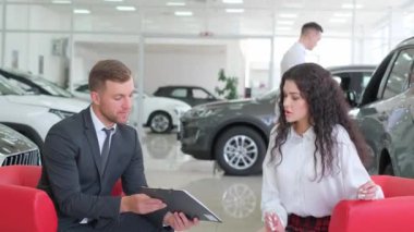 Bir adam araba galerisinde bir kadınla otomotiv tasarımını tartışıyor. Aracın parlak tekerleğine ve lastik özelliklerine doğru hareket ediyor.