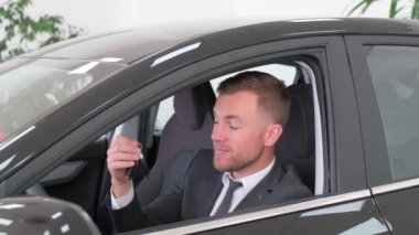 Sivri takım elbiseli ve kravatlı bir adam lüks bir arabada oturmuş telefonunu kontrol ediyor. Araç kapısı ve araç koltuğu gibi otomotiv tasarımı detaylarıyla çevrili.