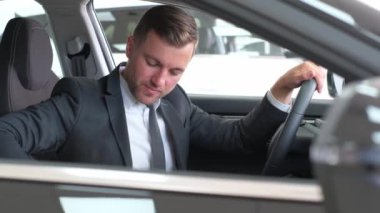 Takım elbiseli bir adam bir araçta oturuyor ve aynası ayarlı. Arabalar gösterişli araba tasarımı ve parlak tekerlekler onun profesyonel jestini bu ulaşım biçiminde yansıtıyor.