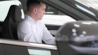 Bir adam, bir motorlu aracın, özellikle de otomotiv aynaları ve araç kapısı ile çevrili bir arabanın sürücü koltuğunda oturmaktadır.