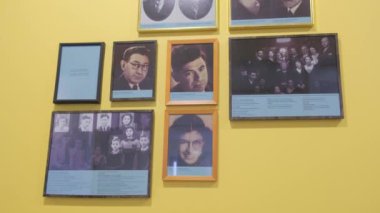 Bir video çerçevelenmiş insanların resimleriyle süslenmiş sarı bir duvar gösteriyor, her birinin kendine özgü bir hikayesi var. Renkli ve nostaljik bir atmosferde çekilen fotoğraflardaki kişisel anıları vurgular.