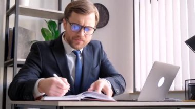 Takım elbiseli ve kravatlı bir işadamı elinde laptop ve kitapla bir masada oturuyor, işine odaklanmış ve meşgul.
