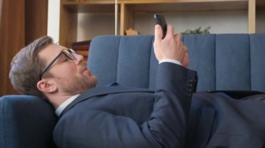 Videoda, takım elbiseli bir adam mavi bir kanepede telefonuna bakıyor, uyuyor ve gözlüklerini çıkarıyor. Rahatlamış ve dinlenirken telefonuna odaklanmış görünüyor.