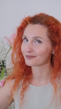 Kırmızı saçlı ve mavi gözlü neşeli bir kadın canlı bir buketin önünde duruyor. Sıcak bir şekilde gülümsüyor, pozitif ve neşe saçıyor.