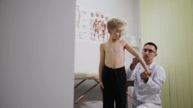 Klinik bir ortamda, genç bir çocuk tıbbi muayeneden geçer. Doktor kolunu ve genel sağlık durumunu değerlendirerek pediatrik bakımı vurguluyor.