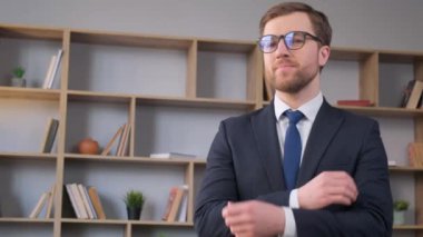 Takım elbiseli ve kravatlı profesyonel bir adam kitaplığın önünde kollarını kavuşturarak duruyor. Zeka ve entelektüellik yayıyor. Güçlü ve otoriter görünüyor.