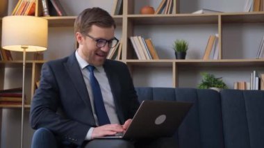 Takım elbiseli ve kravatlı profesyonel bir adam kanepede otururken dizüstü bilgisayarda çalışır, rahat bir ortamda odaklanma ve konsantrasyon gösterir.