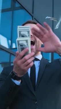 Çılgınca mutlu genç iş adamı cam bir binanın arkasında dikilirken etrafa para saçıyor. Yüksek kazanç. Yüksek maaş. Dikey video. Zengin bir adam.