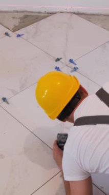 Dikey video. Sarı bir peri masalındaki erkek tamirci yere yeni bir beyaz fayans seriyor. Evdeki modern tadilat. Modern seramik fayanslar