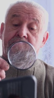 Dikey video. Yaşlı, kır saçlı, sakallı bir adam büyüteçten bakıyor. Yaşlılar. Yaşlılık