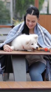Mutlu bir kadın evcil köpeğiyle dışarıda bir masada oturuyor. Beyaz tüylü köpeği olan bir kadın. Mutlu insanlar. Dikey video.