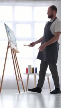 Uzun boylu, erkek bir sanatçı atölyesinde bir resim çiziyor. Beyaz kaplamalı modern sanat stüdyosu. Dikey çekim.