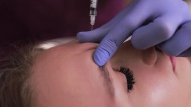 Alnına botoks yapılan bir kadın dış görünüşünü güçlendiriyor. Pratisyenler mavi parmaklarını tenine bastırırken onun mor kirpikleri titriyor. Dikey video