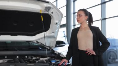 Bir araba galerisinde açık bir arabanın kaputunun yanında takım elbiseli genç bir kadın. Yüksek kalite 4k görüntü