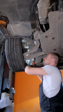 Bir adam yerde bir aracı tamir ediyor, otomotiv lastikleri üzerinde çalışıyor. Çamurluk ve tampon otomobil parçaları da dahil olmak üzere kenar ve otomotiv dış yüzeyine odaklanmış durumda. Dikey video