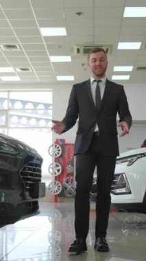 Resmi giyinmiş bir adam, çeşitli araçlarla çevrili bir galeride. Sergilenen arabaların otomotiv tasarımı ve ışıklandırma özelliklerini inceliyor. Dikey video