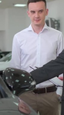 Adam araba galerisinde araba satıcısıyla konuşuyor. Etrafı arabalar, araba tasarımı, kaputlar, aydınlatma, kapılar ve lüks arabalarla çevrili. Dikey video