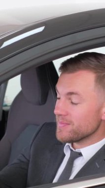 Sivri takım elbiseli ve kravatlı bir adam lüks bir arabada oturmuş telefonunu kontrol ediyor. Araç kapısı ve araç koltuğu gibi otomotiv tasarımı detaylarıyla çevrili. Dikey video