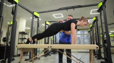 Bir erkek, spor salonundaki bir masada şınav çekerken bir kadına yardım ediyor. İkisi de spor salonunda spor yapıyor.