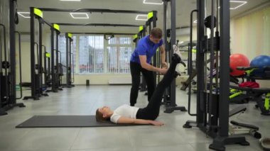 Hareketli bir spor salonunda, bir erkek egzersiz sırasında bir kadına destek olur, bacağını esnek ve dayanıklı gösterir, takım çalışmasını vurgular.