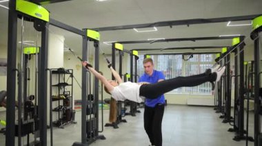 Canlı bir spor salonunda, bir erkek ekipman üzerinde çalışırken bir kadına destek olur, takım çalışmasının ve fitness motivasyonunun pozitif atmosferini vurgular.
