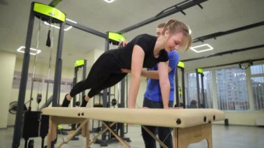 Masada şınav çeken bir kadını destekleyen bir erkek, takım çalışmasını ve kişisel hedefleri doğrultusunda çalışan fitness motivasyonunu ön plana çıkarıyor.