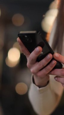 Resmi kıyafetli bir kadın elinde bir cep telefonu tutuyor, baş parmağını cihazın yazı tipini yazmak için kullanıyor. Tırnağı boyalı ve bir elbise giyiyor, bilek aleti ile aksesuar takıyor. Dikey video